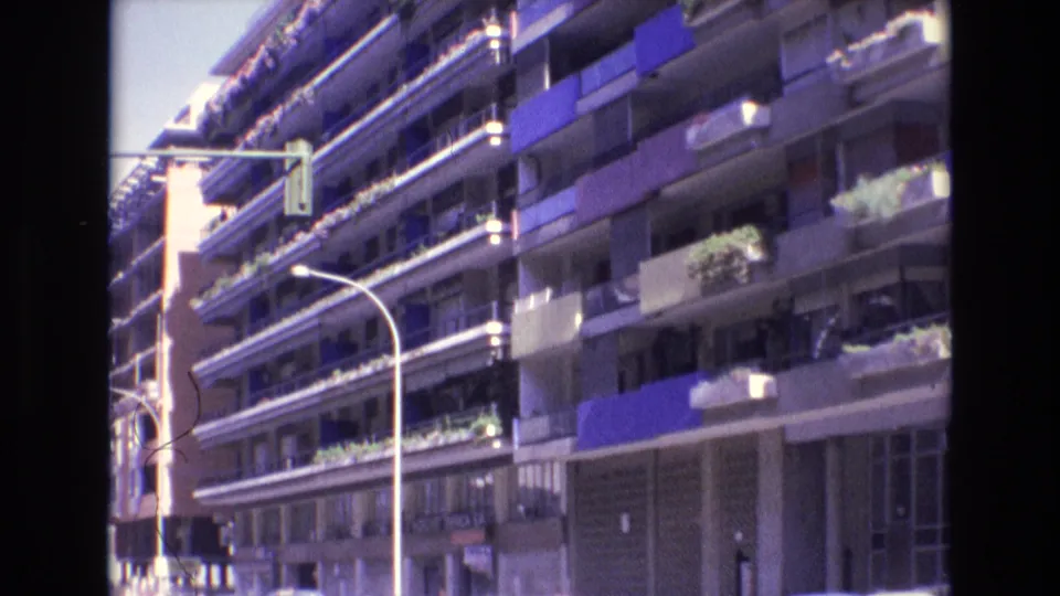 1972 Spain home-movie frame showing a sunlit apartment exterior with repeated balconies, blue awnings, planted balcony edges, a curved street lamp, and a traffic light at the street corner.