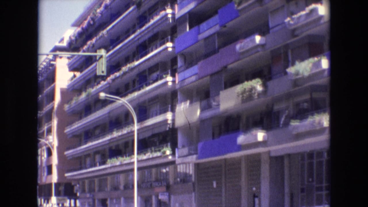 1972 Spain street view of a multi-story apartment building with balcony plants, blue awnings, shopfront level, and a curved streetlight in the foreground.