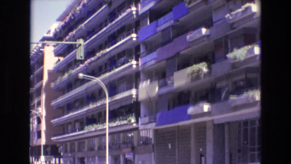 1972 Spain street view of a multi-story apartment building with balcony plants, blue awnings, shopfront level, and a curved streetlight in the foreground.