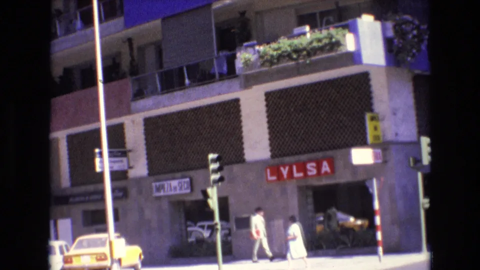 1972 Spain downtown sidewalk view with the LYLSA sign, traffic lights, two pedestrians, a yellow car, and apartment balconies above the street level.