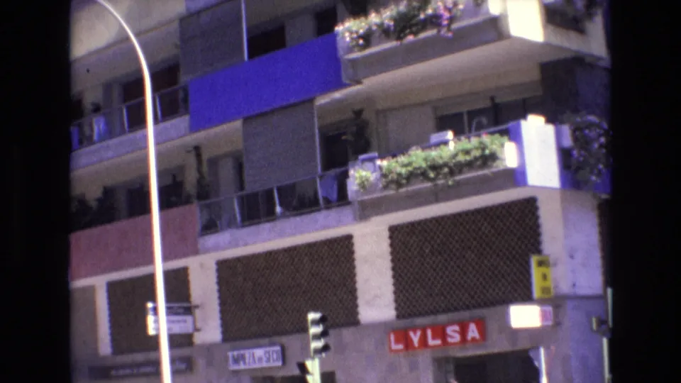 Spain 1972 urban facade with planted balconies, blue awning panels, traffic signals, a tall street pole, and the red LYLSA sign at street level.