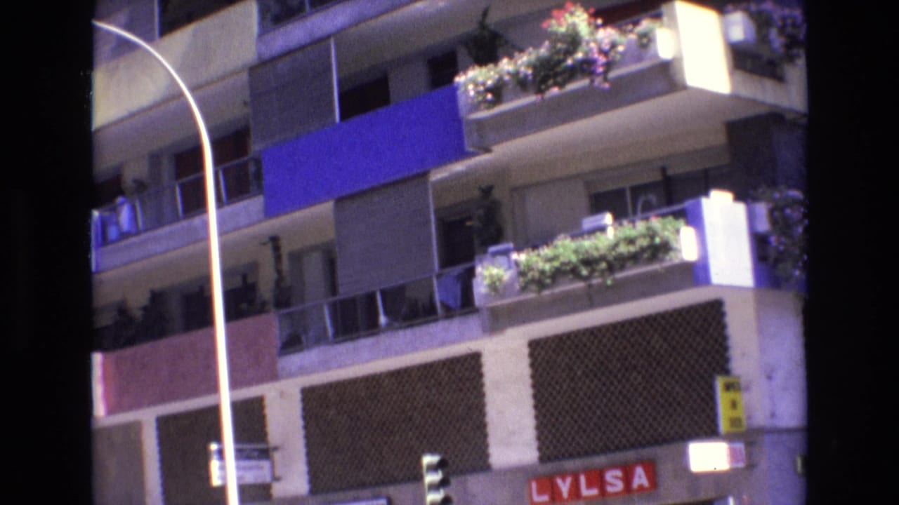 1972 Spain 8mm frame showing a residential apartment facade with stacked balconies, potted flowers, a blue awning panel, a curved street lamp, and partial ground-floor signage.