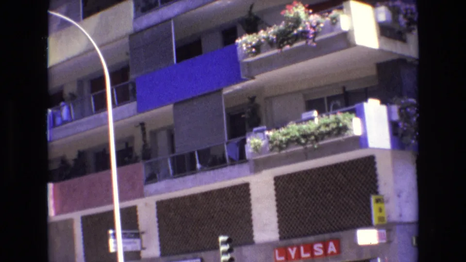 1972 Spain 8mm frame showing a residential apartment facade with stacked balconies, potted flowers, a blue awning panel, a curved street lamp, and partial ground-floor signage.
