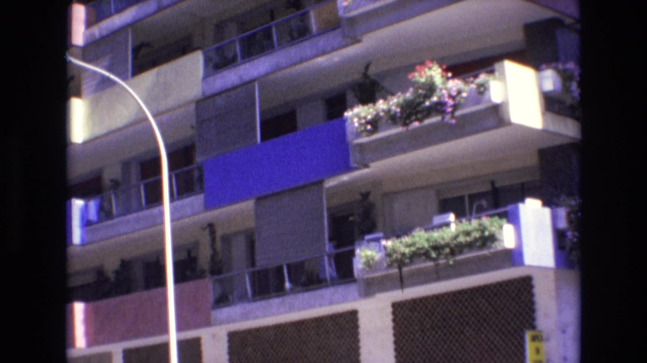Upper apartment balconies with flower planters, blue awning, street lamp, and facade panels.