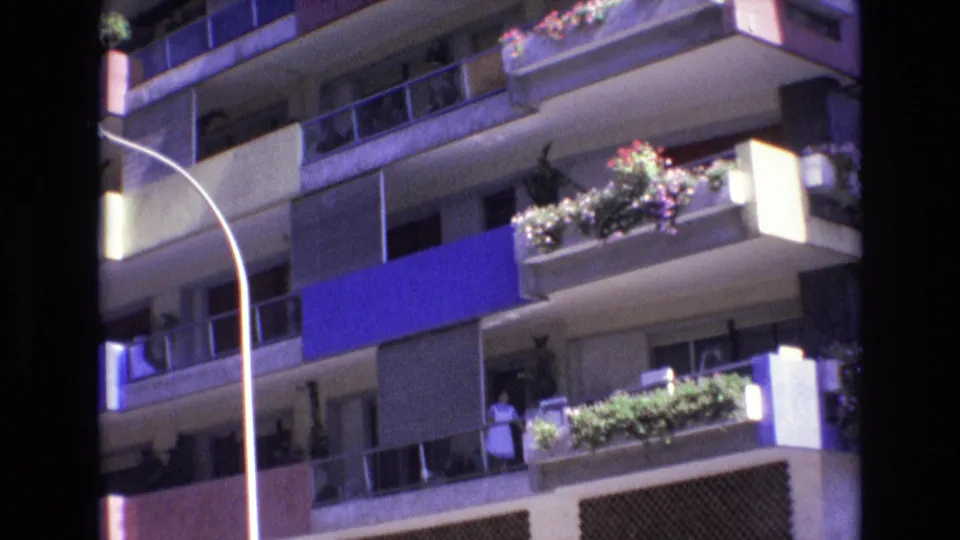 1972 Spain 8mm frame showing apartment balconies with a blue awning, potted plants, a curved street lamp, and a small visible person near a balcony opening.