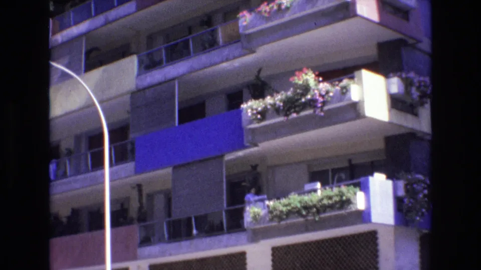 1972 Spain 8mm frame of a residential apartment facade with stacked balconies, flower boxes, a blue awning panel, curved street lamp, and lower patterned facade panels.
