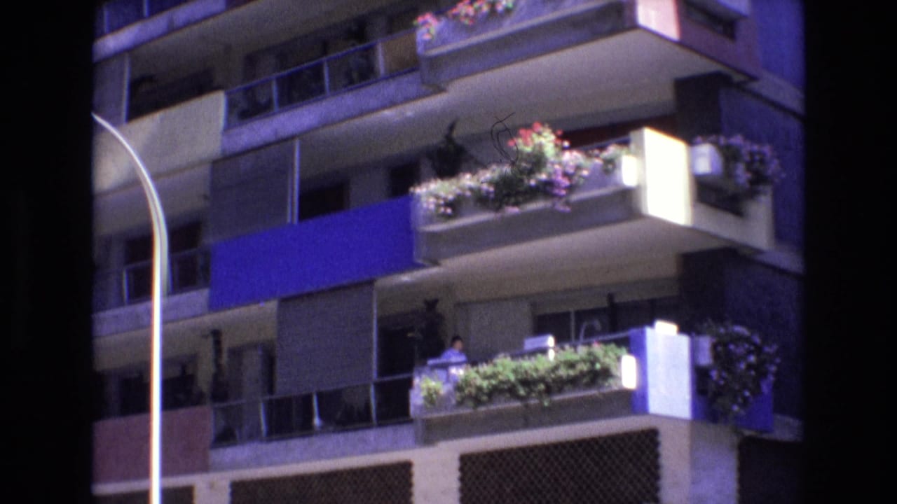 1972 Spain 8mm frame showing a multi-story apartment facade with planted balconies, blue accent panels, traffic lights, a curved street lamp, and a red LYLSA sign at street level.