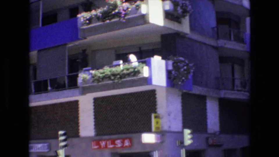 1972 Spain 8mm frame of an apartment building corner with planted balconies, blue panels, patterned facade sections, traffic signals, and a visible LYLSA sign below.