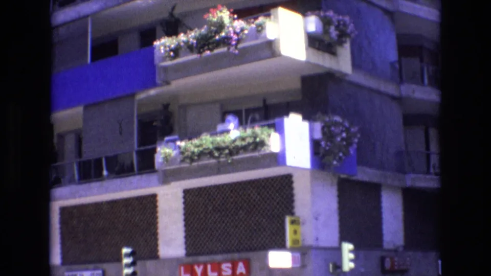 1972 Spain 8mm frame showing a person in light-colored clothing leaning or standing on a planted apartment balcony, with blue facade panels, traffic lights, and LYLSA signage below.
