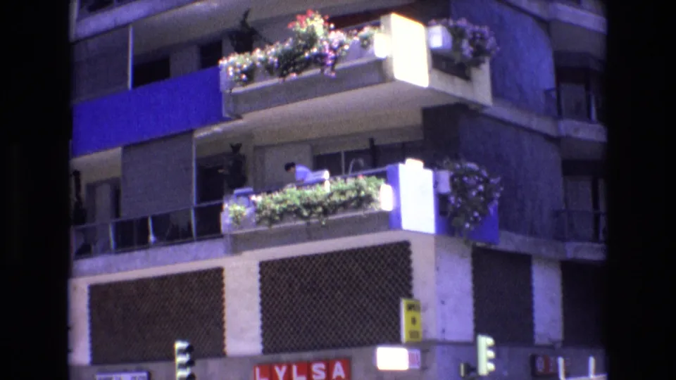 1972 Spain 8mm frame showing a person on a planted balcony of a multi-story apartment building, with blue architectural accents, patterned facade panels, traffic lights, and partial street signage.