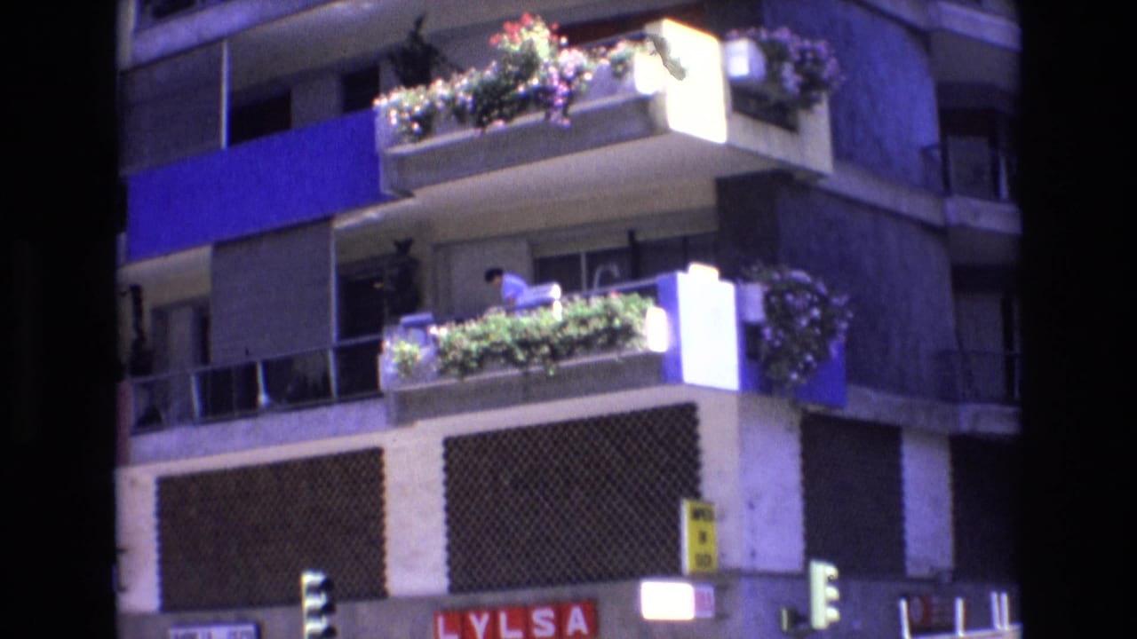 1972 Spain 8mm frame showing a multi-story apartment facade with blue panels, balcony flowers, a person leaning near the railing, and street-level lights below.