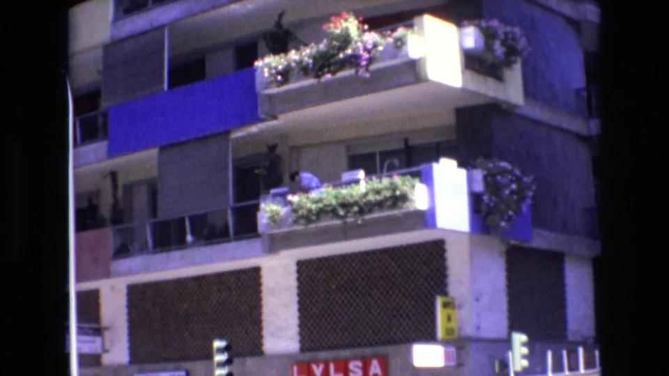 1972 Spain 8mm frame showing stacked balconies with flowers and greenery, a person tending plants, blue facade panels, and partial street-level signage.