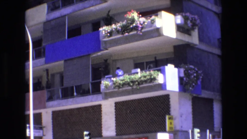 1972 Spain 8mm frame of a residential apartment corner with illuminated balconies, planted railings, a person in a light shirt, and traffic lights near the street.