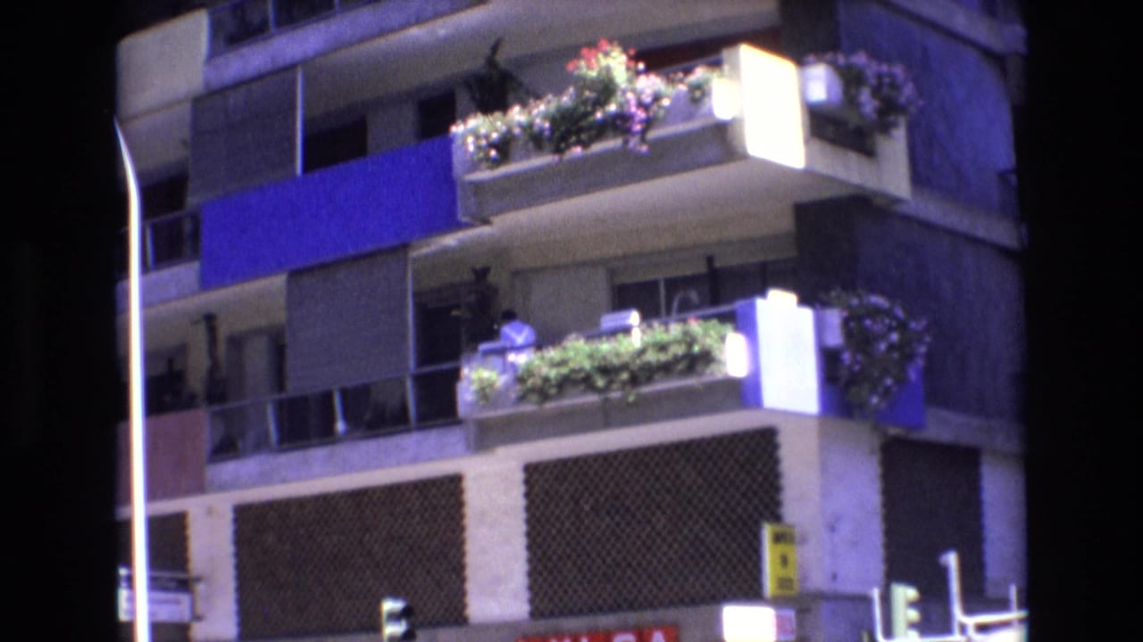 1972 Spain 8mm frame showing a sunlit apartment facade with balcony planters and shadowed street edges.