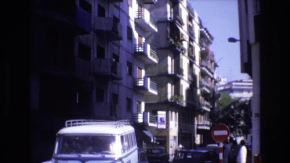 1972 Spain 8mm frame with a white van in the foreground, apartment balconies above, parked cars, pedestrians, and a red do-not-enter sign at the curb.