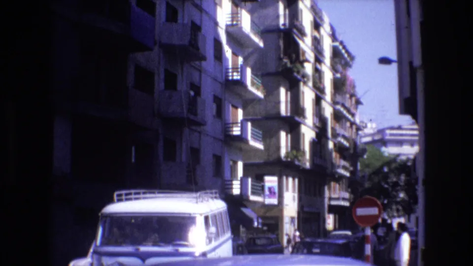 1972 Spain 8mm frame showing the same apartment-lined street with a white van, balcony planters, parked cars, distant pedestrians, and strong blue-purple film color shift.