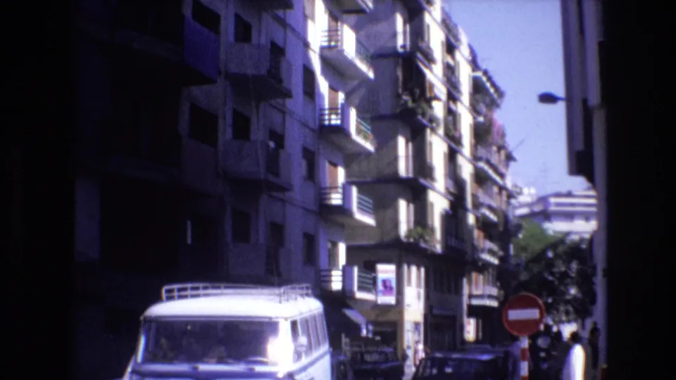 A white van with a roof rack moves along a Spanish city street in the supplied 1972 footage, framed by tall apartment buildings with balconies and a red no-entry sign.