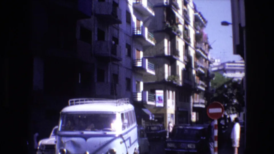 Traffic moves through a bright urban street in Spain, with a white van in the foreground, parked cars, apartment balconies, and a red no-entry sign visible near the curb.