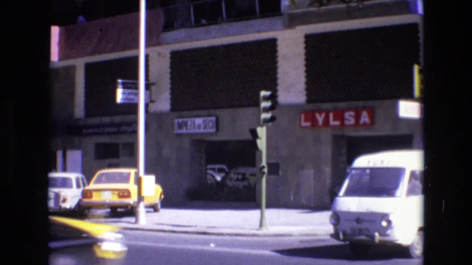 A yellow taxi-like car and a white delivery van marked with lettering move past storefronts and traffic lights in the supplied 1972 Spain street footage.