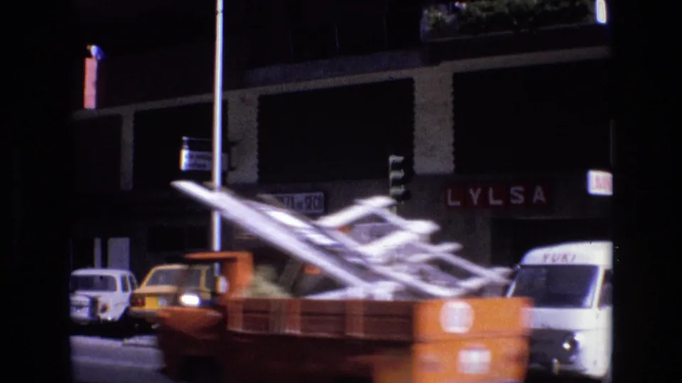 An orange truck carrying a large white ladder crosses the frame with motion blur, passing storefront signage, parked cars, and a white delivery van on a Spanish city street.