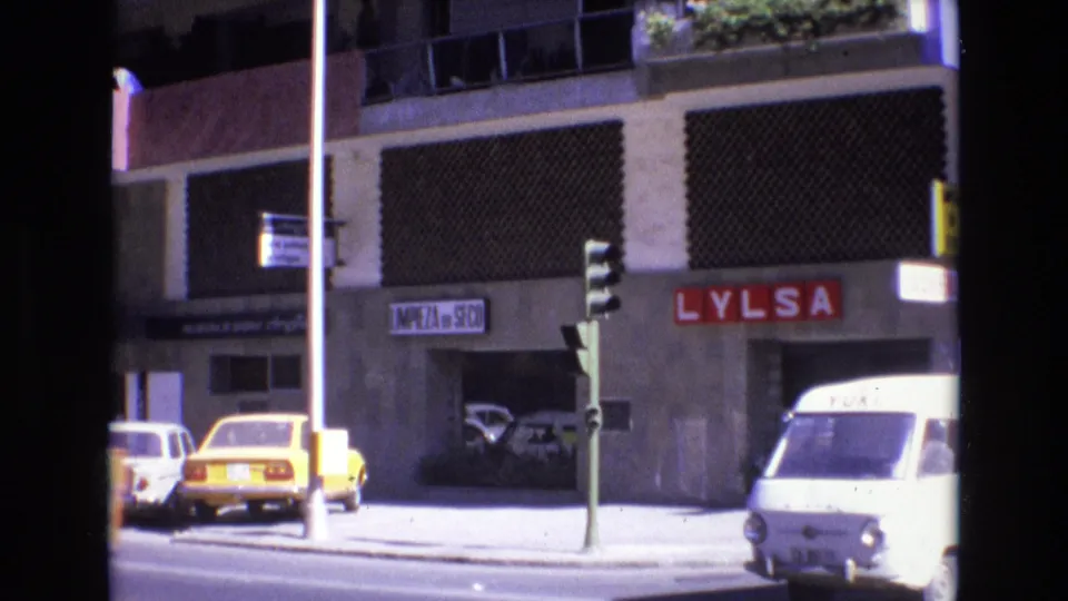 1972 Spain 8mm street view with compact cars, storefront signage including a red sign, a traffic light, and a white van near the curb.