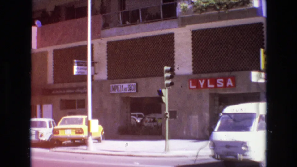 1972 Spain street frame showing storefronts, visible commercial signage, parked cars, a traffic signal, and a white van in bright daylight.