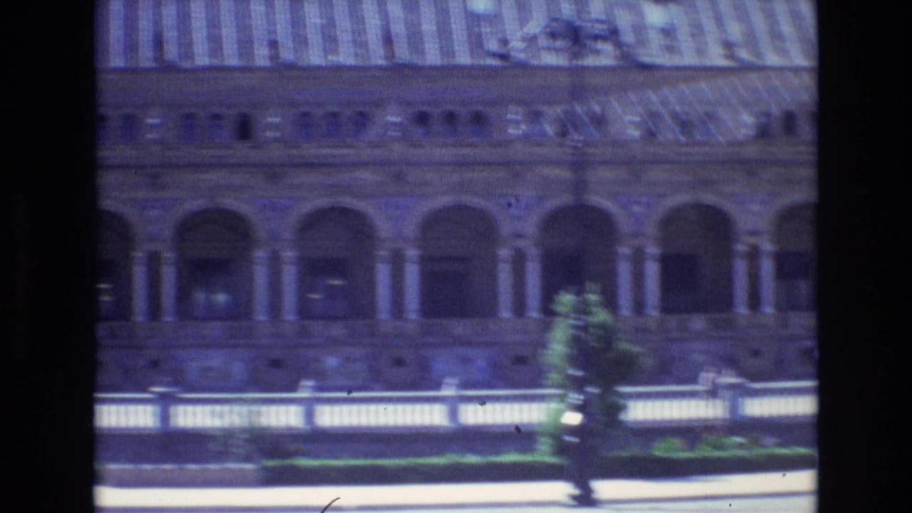 A large Spanish building exterior appears from a moving 1972 home-movie viewpoint, showing arched colonnades, a striped roof, and a pale balustrade in front.
