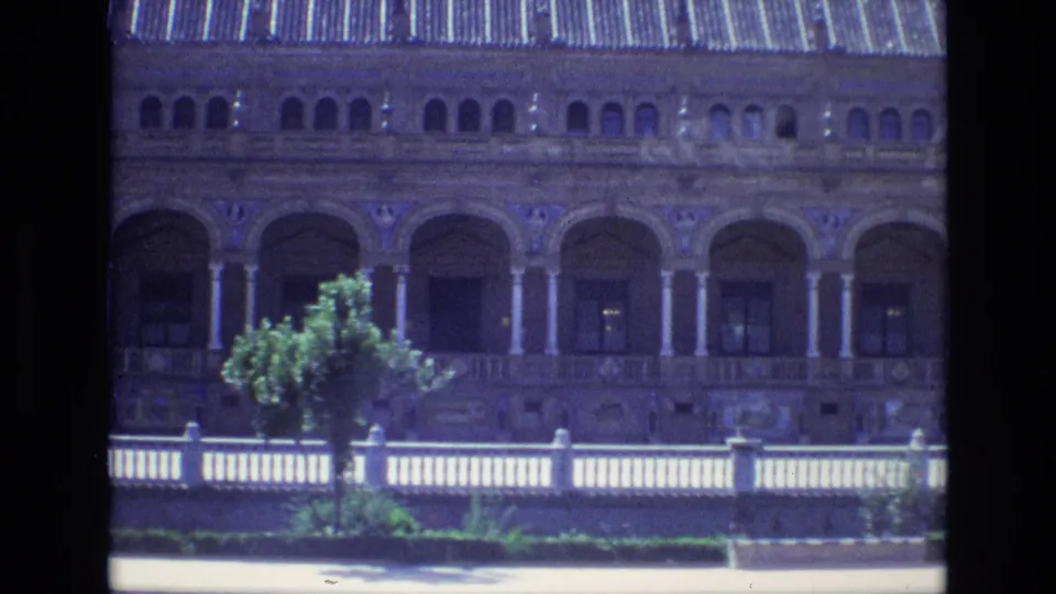 The 1972 Spain travel footage holds on a historic-looking facade with rows of arches, a striped roofline, a pale balustrade, and a small tree in the foreground.