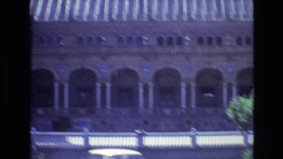 A softer moving frame from the Spain 1972 home movie shows the same arched building exterior with striped roof detail, foreground balustrade, and a partial tree at the edge.