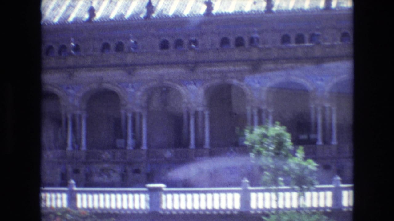 1972 8mm home-movie view of Plaza de Espana in Seville, showing arched colonnades, tiled roofline, white railing, canal edge, and a tree in the foreground.