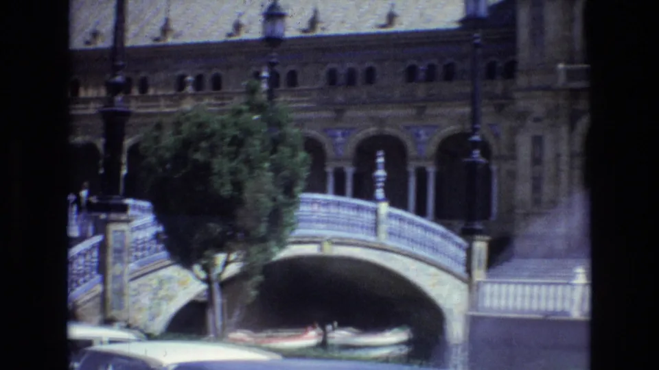 1972 travel-film frame at Plaza de Espana in Seville with a curved bridge over the canal, a foreground tree, lampposts, and ornate arches behind it.