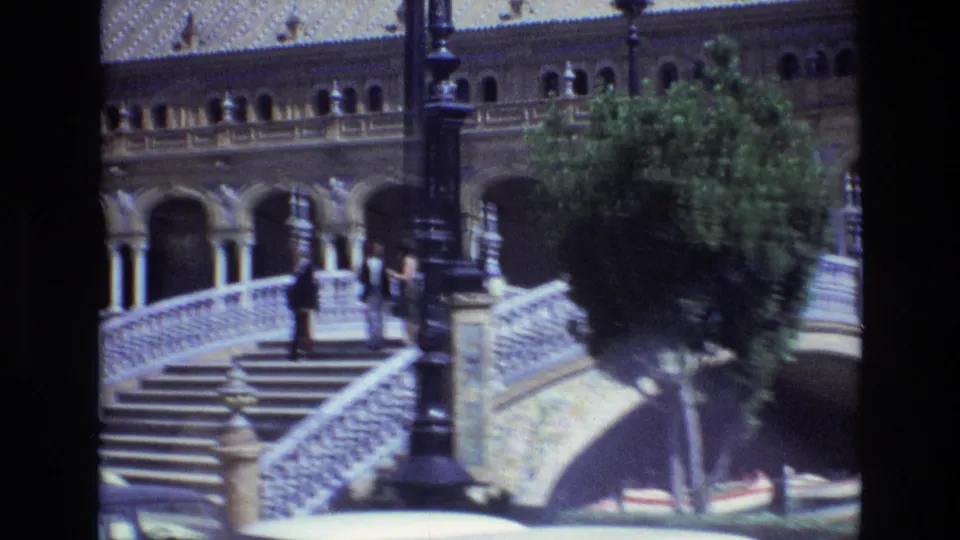 1972 8mm view of a Plaza de Espana bridge stairway in Seville, with white railings, a tall lamppost, arched colonnades, a tree, and distant visitors.