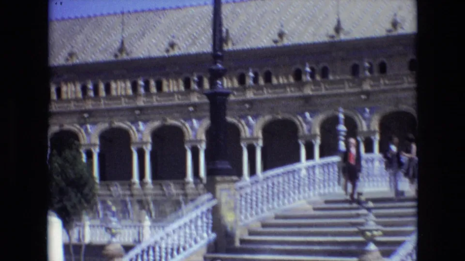 1972 home-movie frame from Plaza de Espana in Seville showing bridge steps, a white balustrade, patterned roof tiles, arched colonnades, and several distant visitors.