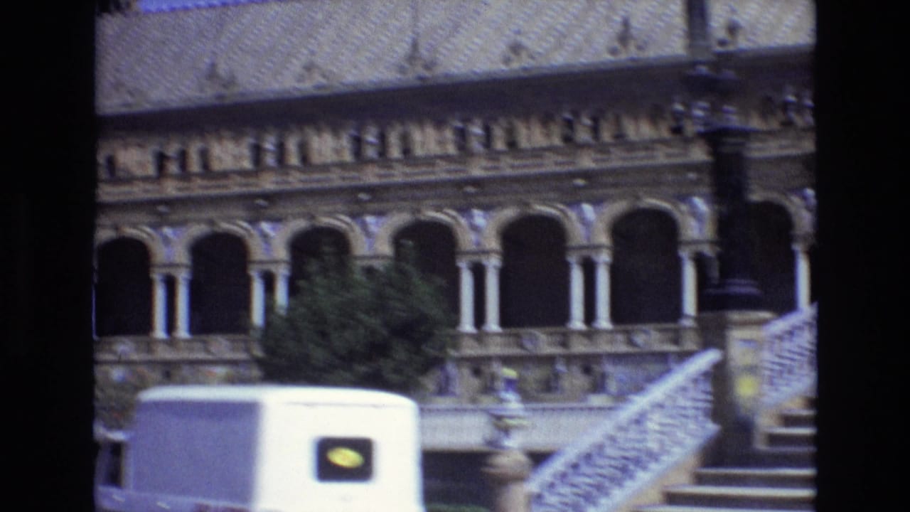 1972 Spain home-movie frame showing a grand historic facade with arched colonnades, roof detail, trees, and a white van passing in the foreground.