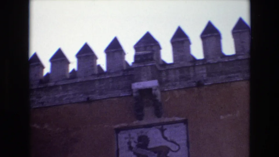 1972 Spain home-movie frame showing a fortress-like crenellated wall, pointed battlements, brick or stucco surface, and a heraldic emblem mounted below.