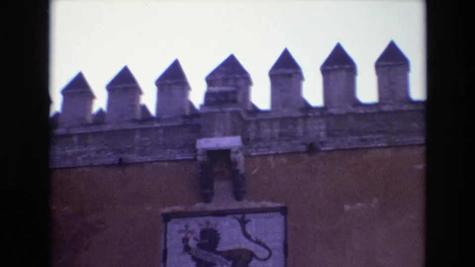 Later 1972 Spain frame showing the same fortress-like wall with pointed battlements, a central mounted emblem, bright sky, and visible film gate edges.