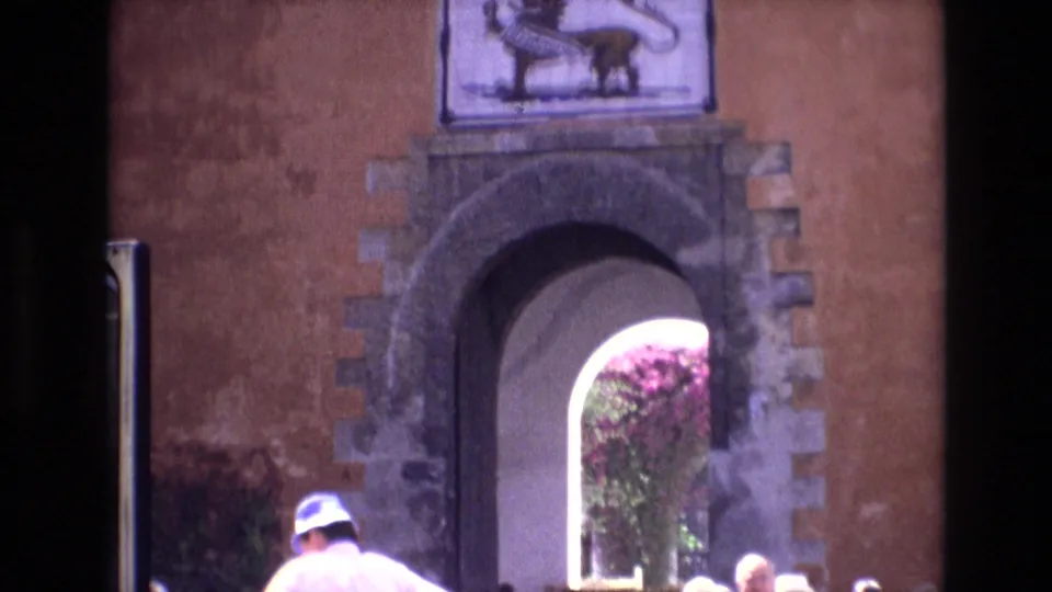 1972 Spain 8mm frame showing tourists at the base of a historic arched entrance, with a crowned lion panel above and vegetation visible through the opening.