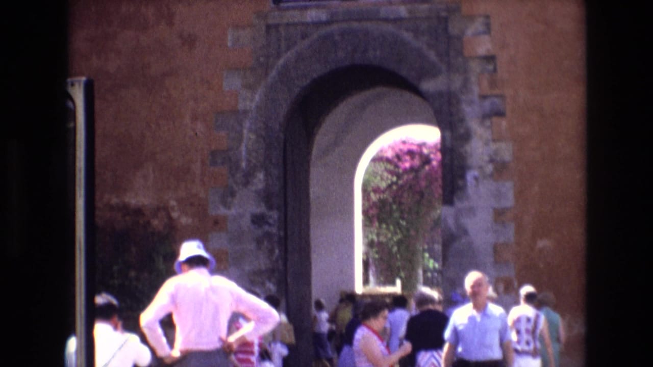 1972 Spain home-movie frame showing tourists in period clothing near a large arched entrance set into a weathered wall.