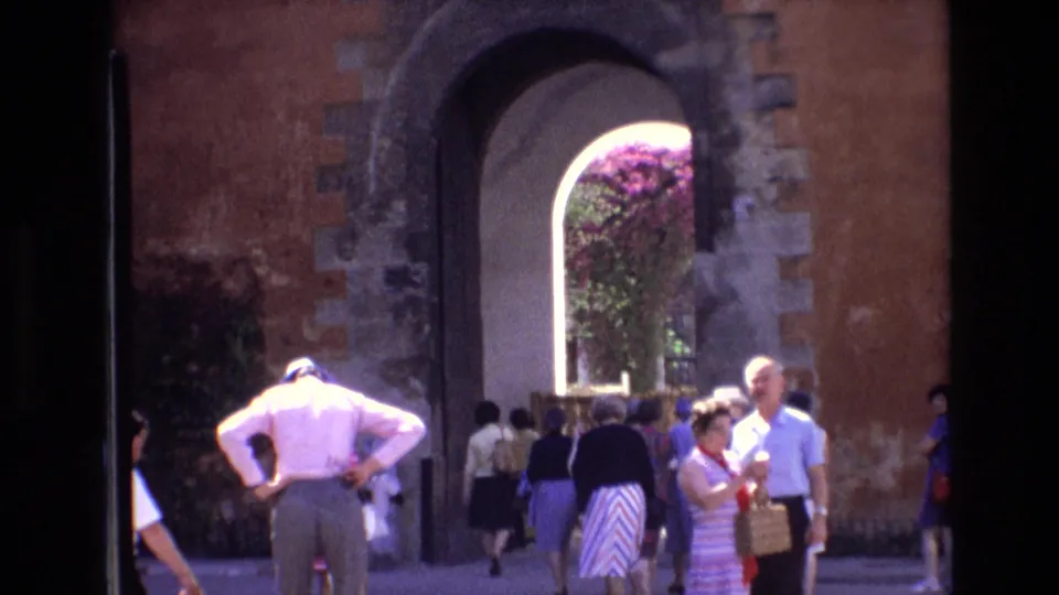 1972 Spain travel footage frame with tourists moving around a tall arched doorway and flowering greenery beyond the opening.