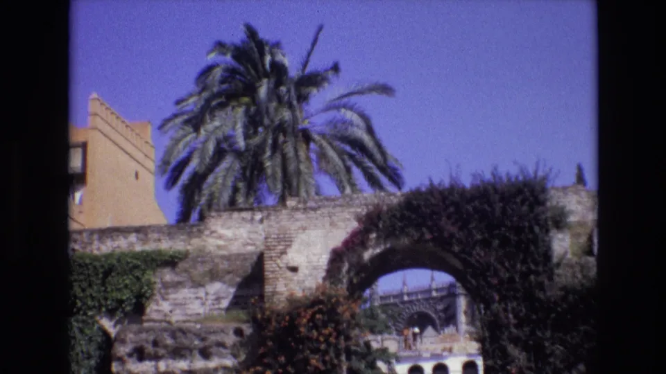 1972 Spain home-movie frame showing a palm tree, ivy-covered stone arch, and distant building details under a clear blue sky.