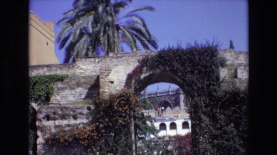 1972 Spain travel frame with an ivy-covered stone arch, palm tree, flowering growth, and a distant building visible through the opening.