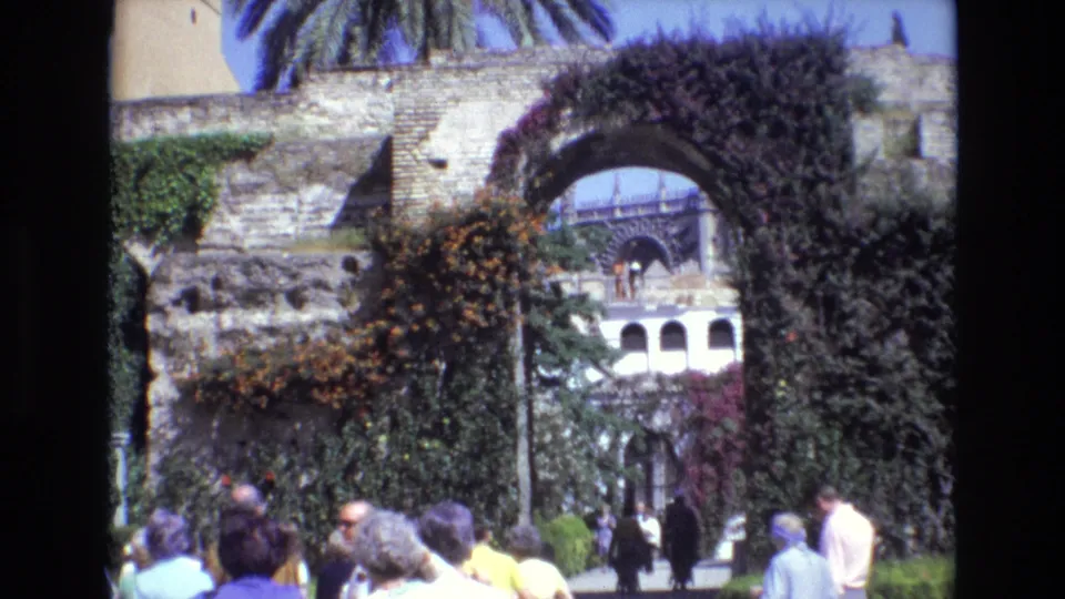 1972 Spain home-movie frame showing a larger group of tourists in period clothing beneath a tall stone arch covered with vines and flowers, with palm fronds against the sky.