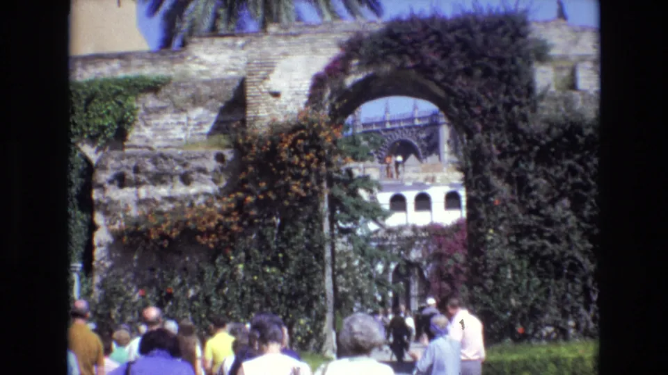 Final 1972 Spain 8mm frame showing tourists in the foreground, a greenery-covered stone arch and wall, palm fronds overhead, and a pale arched building seen through the opening.