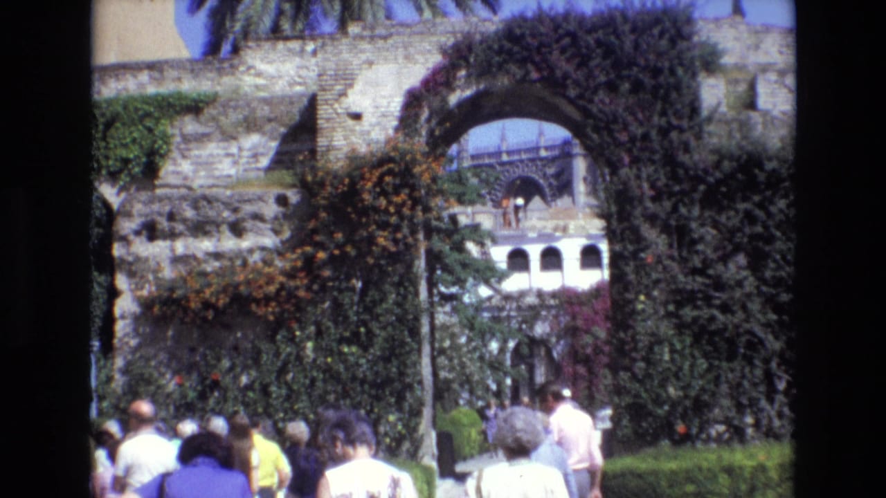 1972 Spain home-movie frame showing visitors passing through a vine-covered stone arch toward a courtyard with historic architecture beyond.