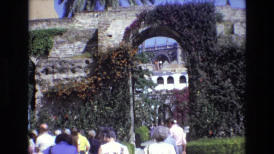 1972 Spain home-movie frame showing visitors passing through a vine-covered stone arch toward a courtyard with historic architecture beyond.