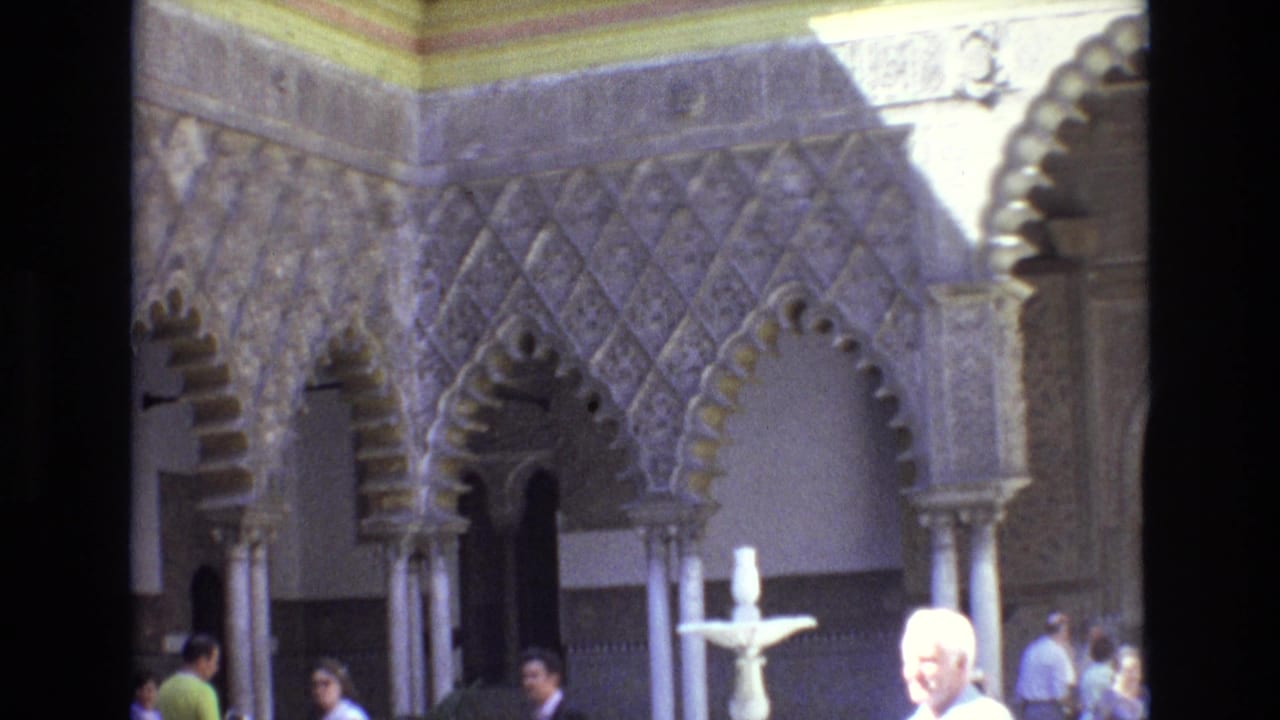 Ornate courtyard wall with carved arches, columns, a fountain, and visitors.