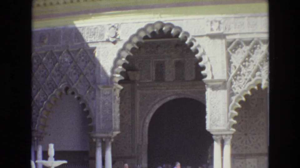 1972 Spain frame focused on a large arched doorway, decorative wall carving, slender columns, a small fountain, and visitors at the bottom of the courtyard view.