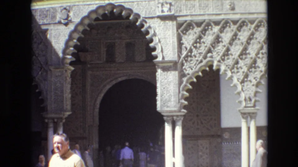 1972 Spain frame showing a decorative arched facade with carved stonework and a briefly visible adult man in a yellow shirt near the lower foreground.