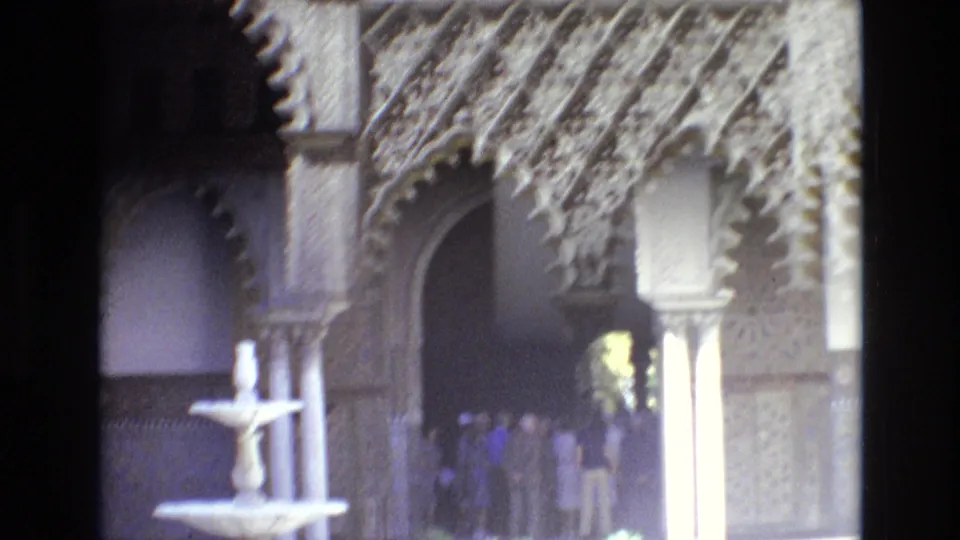 Spain 1972 frame with a small tiered fountain in front of Moorish-style arches and several visitors near a dark passageway.