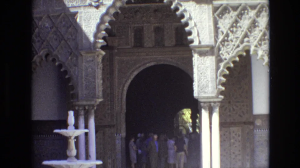 Spain 1972 frame showing visitors gathered in shade beside a small fountain, with ornate Moorish-style arches and patterned courtyard walls around them.